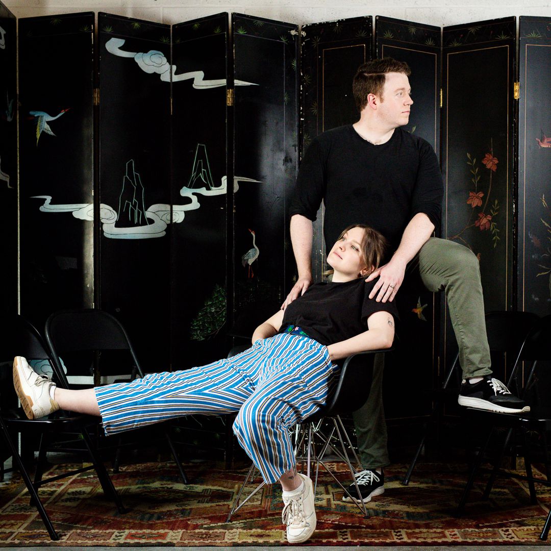 Promo image of Bigger, with Brennan Lee Mulligan and Izzy Roland making silly poses in front of a decorated screen