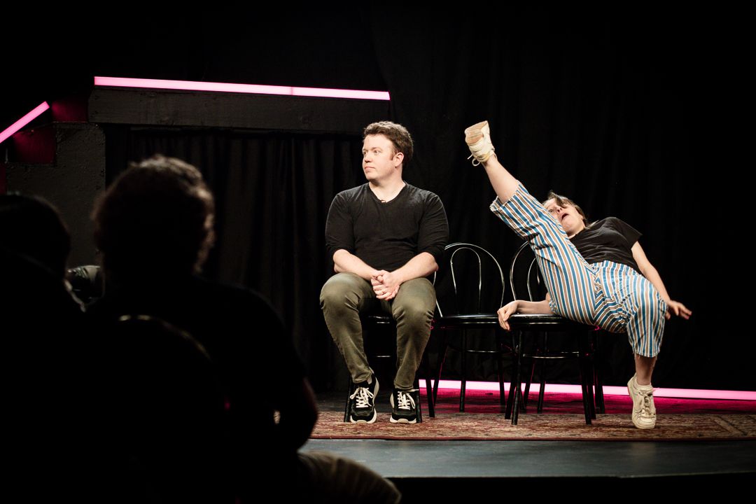 Brennan Lee Mulligan and Izzy Roland doing improv on stage in Bigger. They both sit in chairs, but Izzy kicks her right leg up dramatically