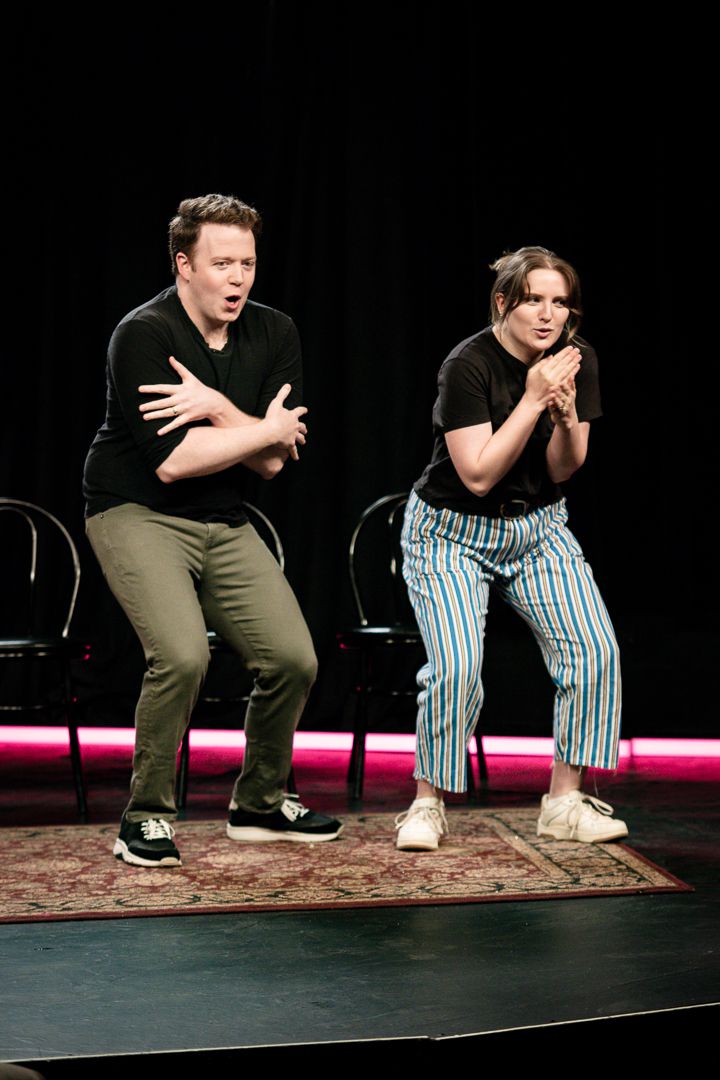 Brennan Lee Mulligan and Izzy Roland both making “I’m cold” gestures on stage in Bigger