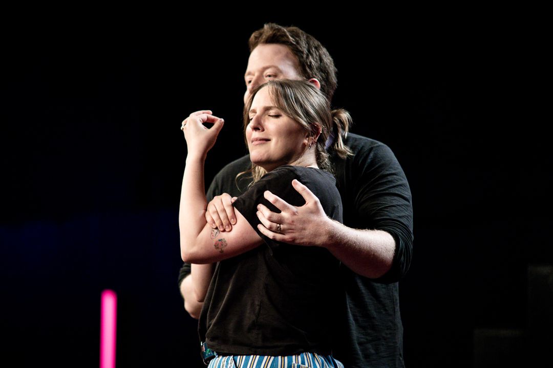 Brennan Lee Mulligan and Izzy Roland doing improv together in Bigger, with Brennan’s arms around Izzy and Izzy’s eyes closed making a gesture