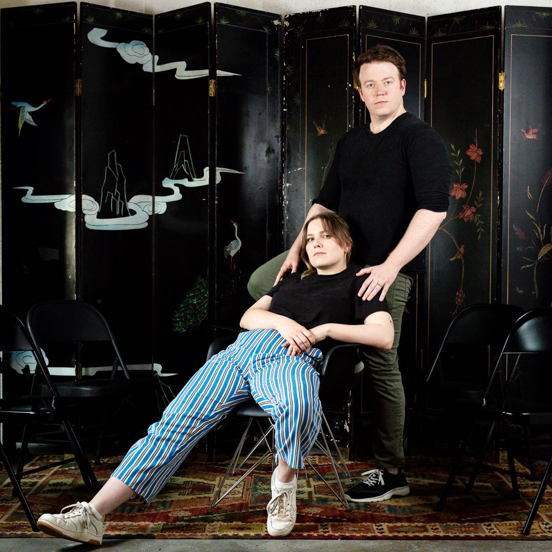 Brennan Lee Mulligan, standing, and Isabella Roland, leaning on a chair, pose in front of a decorated black screen in promo art for Bigger