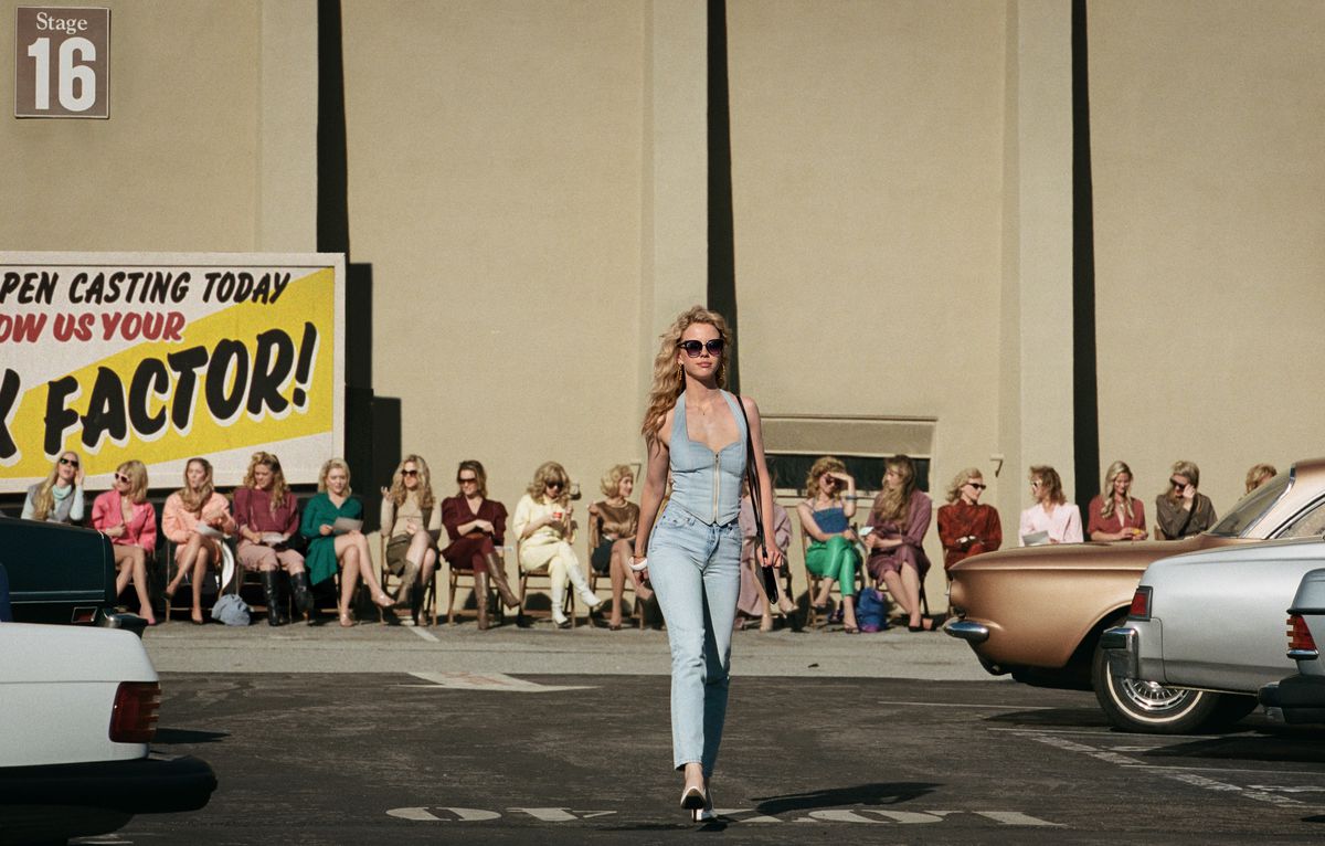 Maxine (Mia Goth) struts across a Hollywood parking lot outside a movie soundstage, with a row of other auditioners lined up in chairs behind her, in Maxxxine 