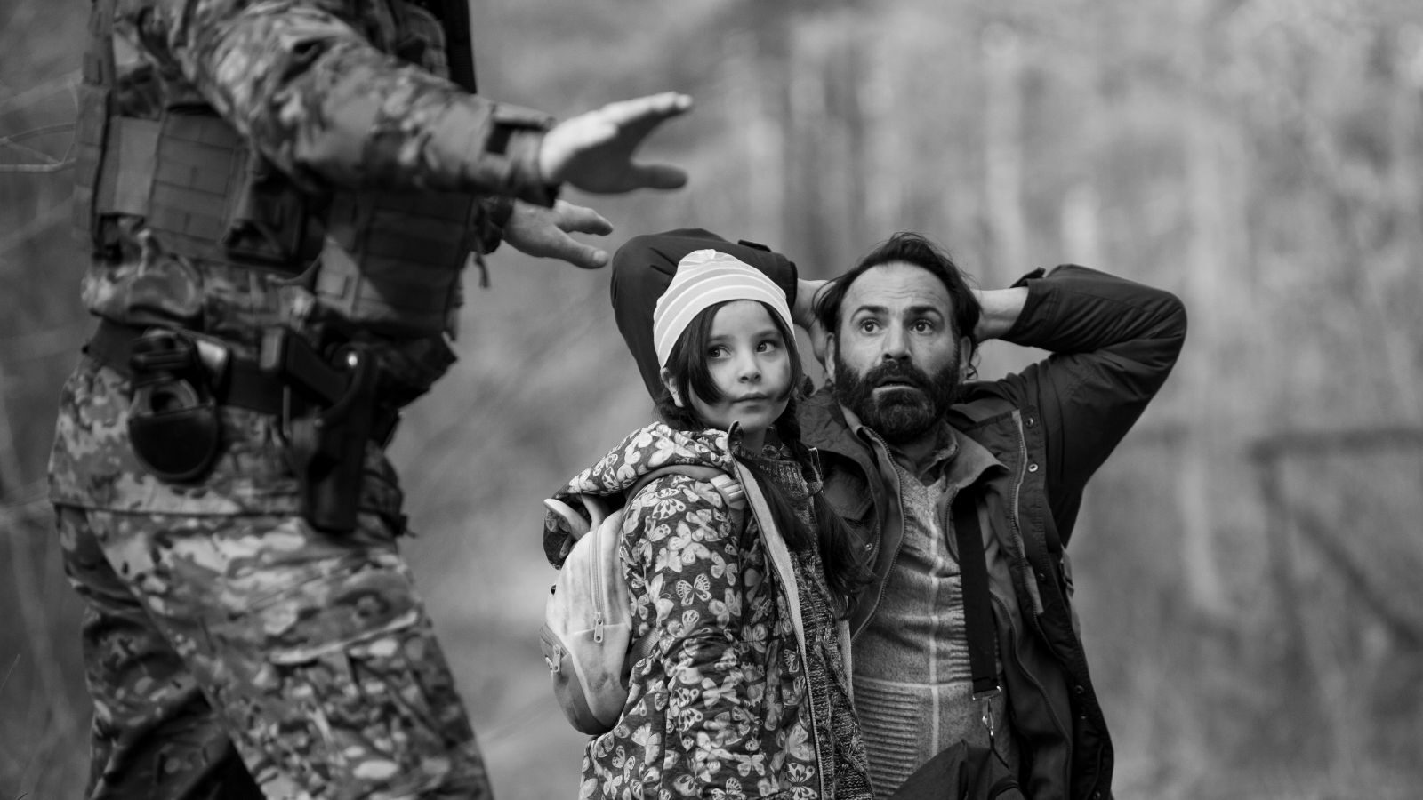 A black and white medium shot of a man on his knees with his hands behind his head next to a child in a winter coat as a soldier in fatigues stands in the foreground with his arms spread in Green Border.