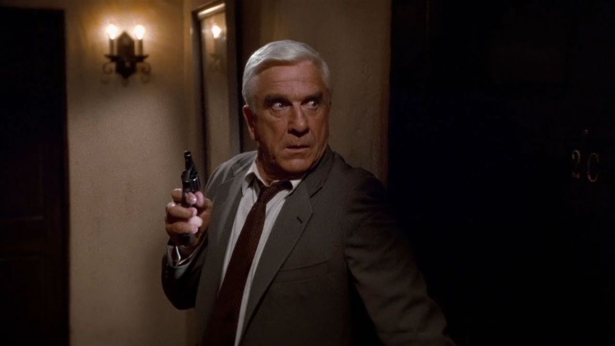 A man in a brown suit holding a small pistol stands next to a wall in The Naked Gun.