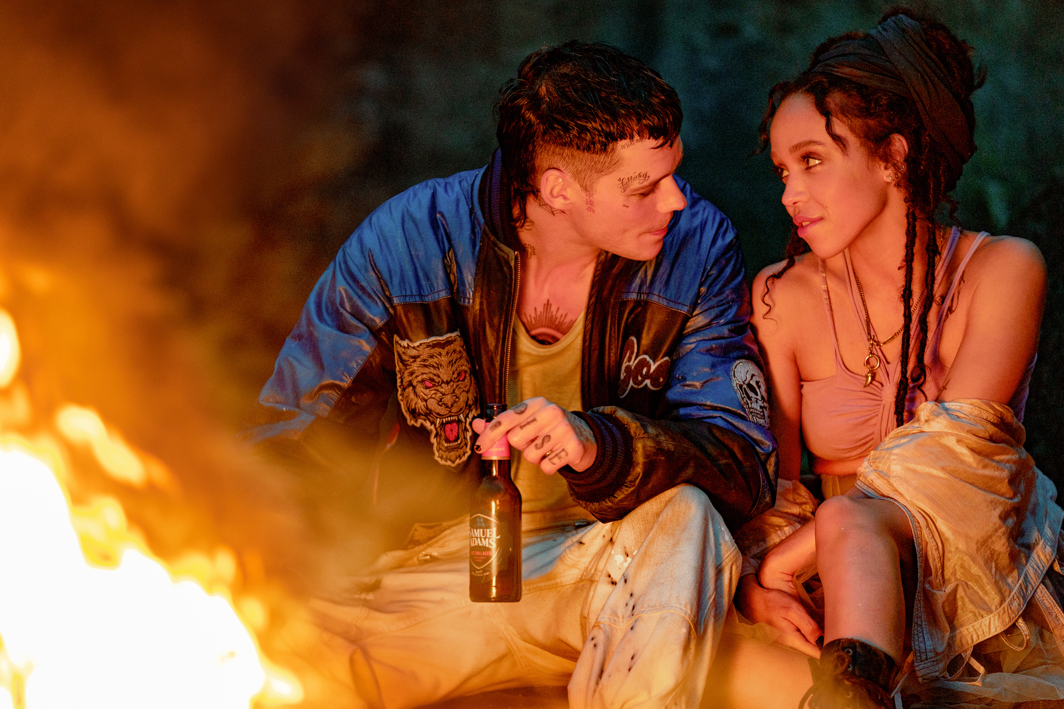 Bill Skarsgård and FKA twigs sit by an outdoor fire in The Crow. Skarsgård holds a beer in his left hand, and they look at each other longingly.