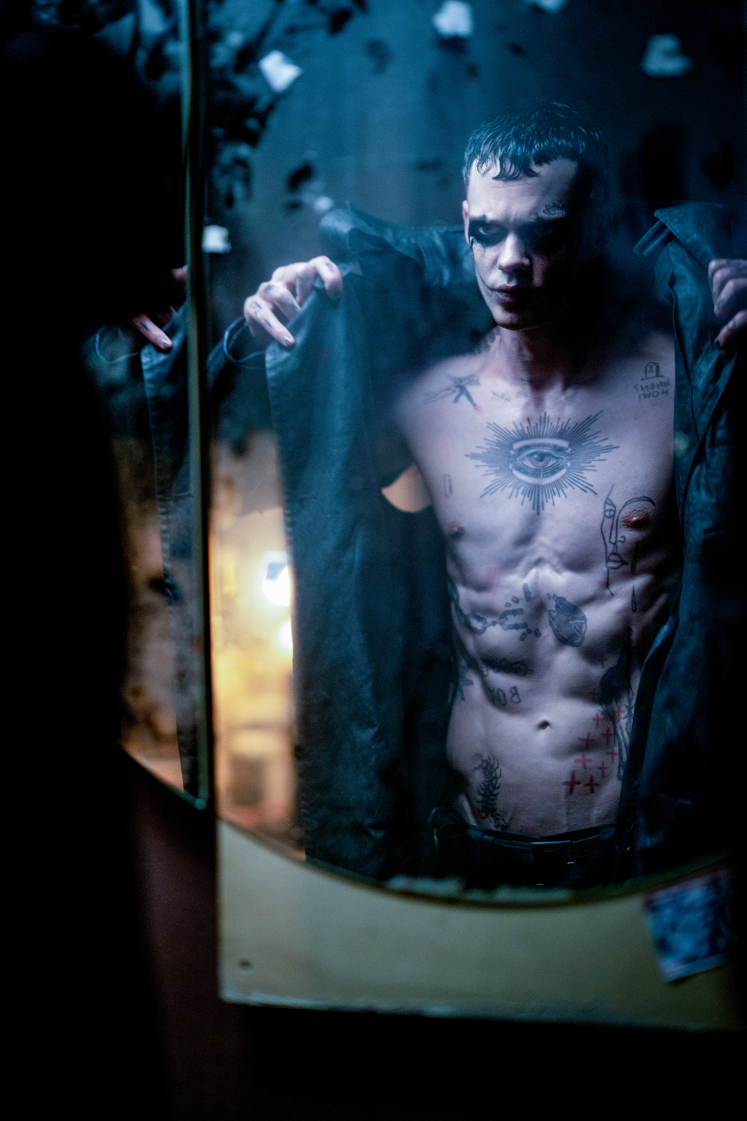 Bill Skarsgård in full Crow get up in The Crow. His jacket is open to show his shirtless torso, which is covered in tattoos. He has heavy black makeup on around his eyes.