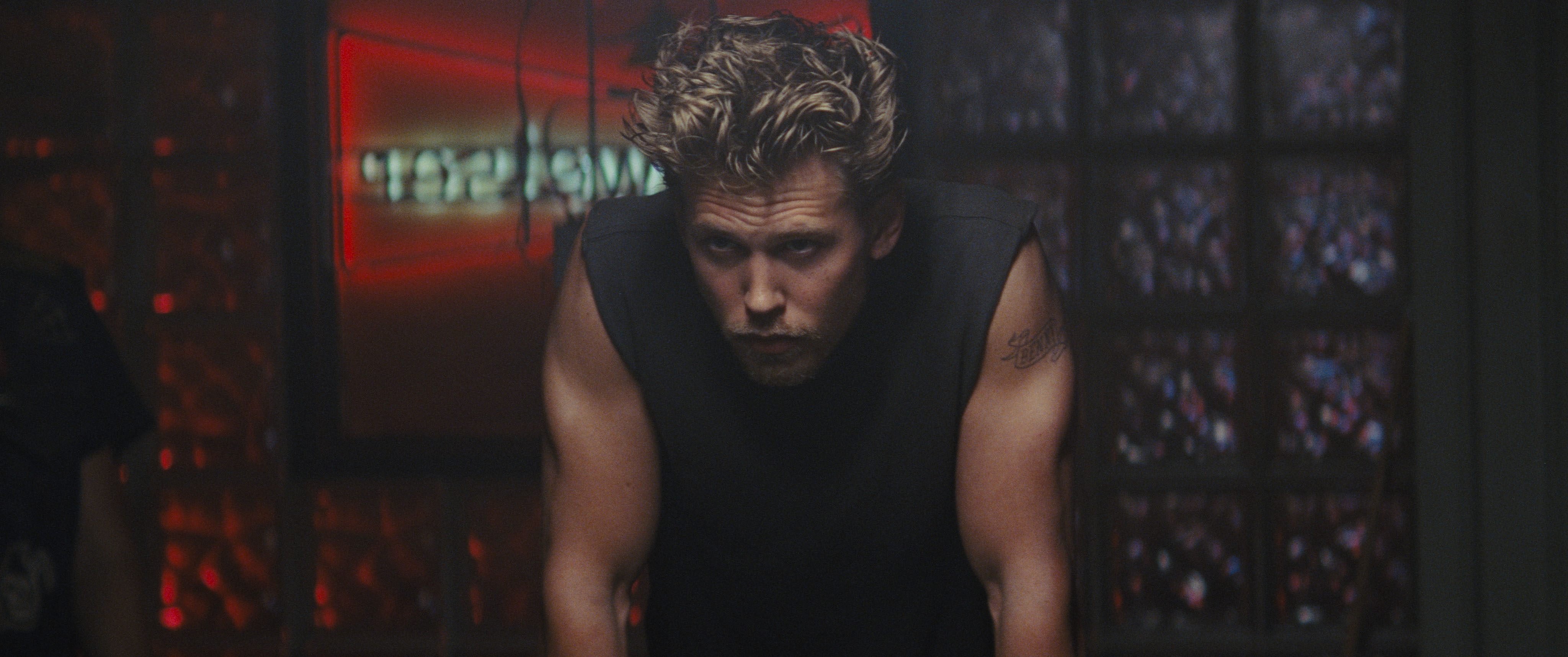 Austin Butler, with mussed-up hair, wearing a black sleeveless top, leans forward in a moody way in The Bikeriders