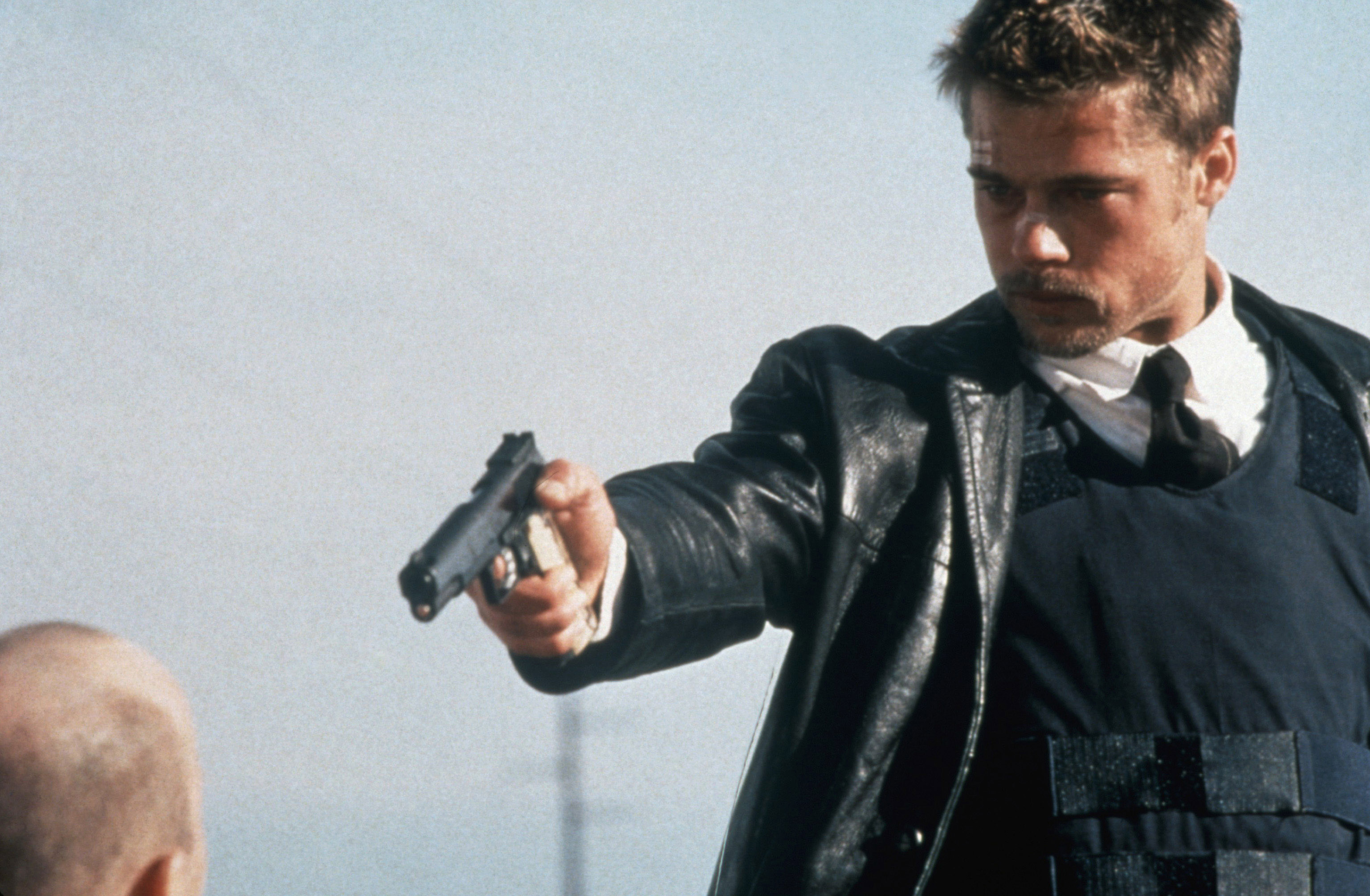 Detective David Mills (Brad Pitt), in bulletproof vest and leather jacket, points his handgun at the shaved-to-the-skin head of John Doe (Kevin Spacey, seen from the back and mostly offscreen) in David Fincher’s Seven