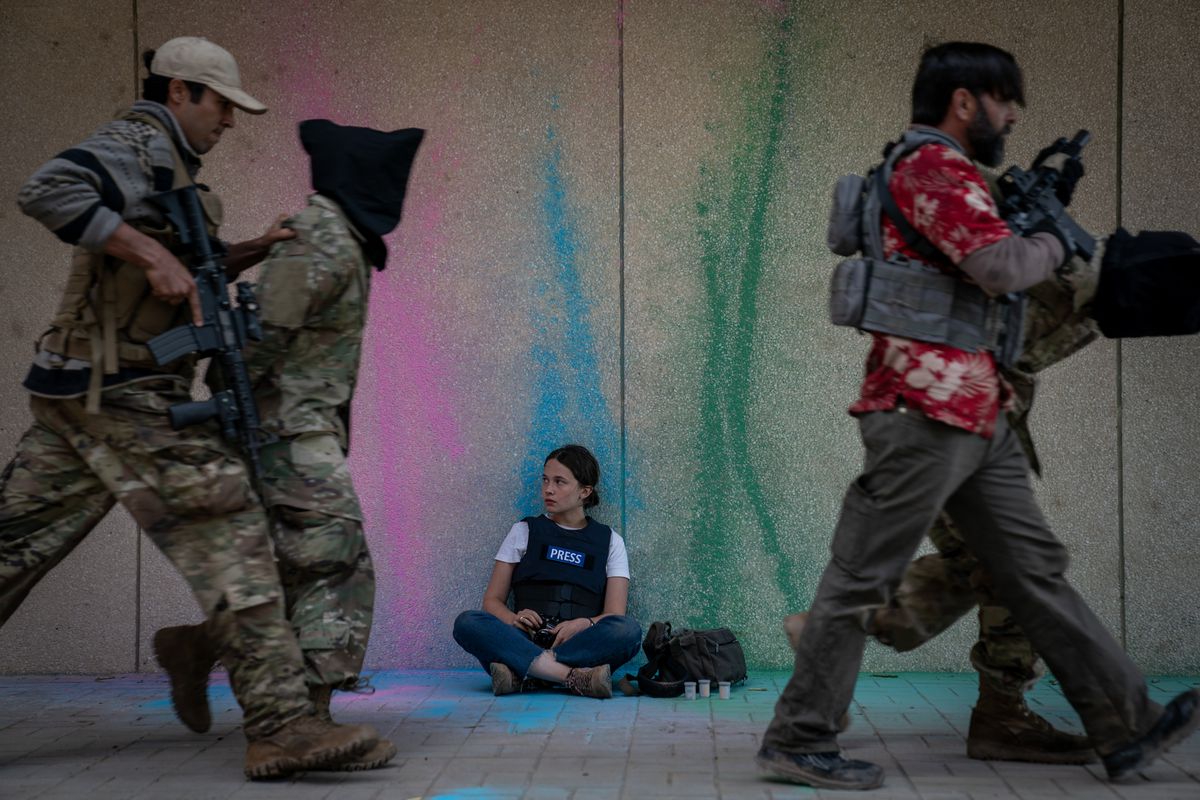 Photojournalist Jessie (Cailee Spaeny) sits with her camera against an exterior wall spattered with pastel slashes of paint as gun-wielding soldiers march by, leading two other soldiers with bags over their heads in Alex Garland’s Civil War