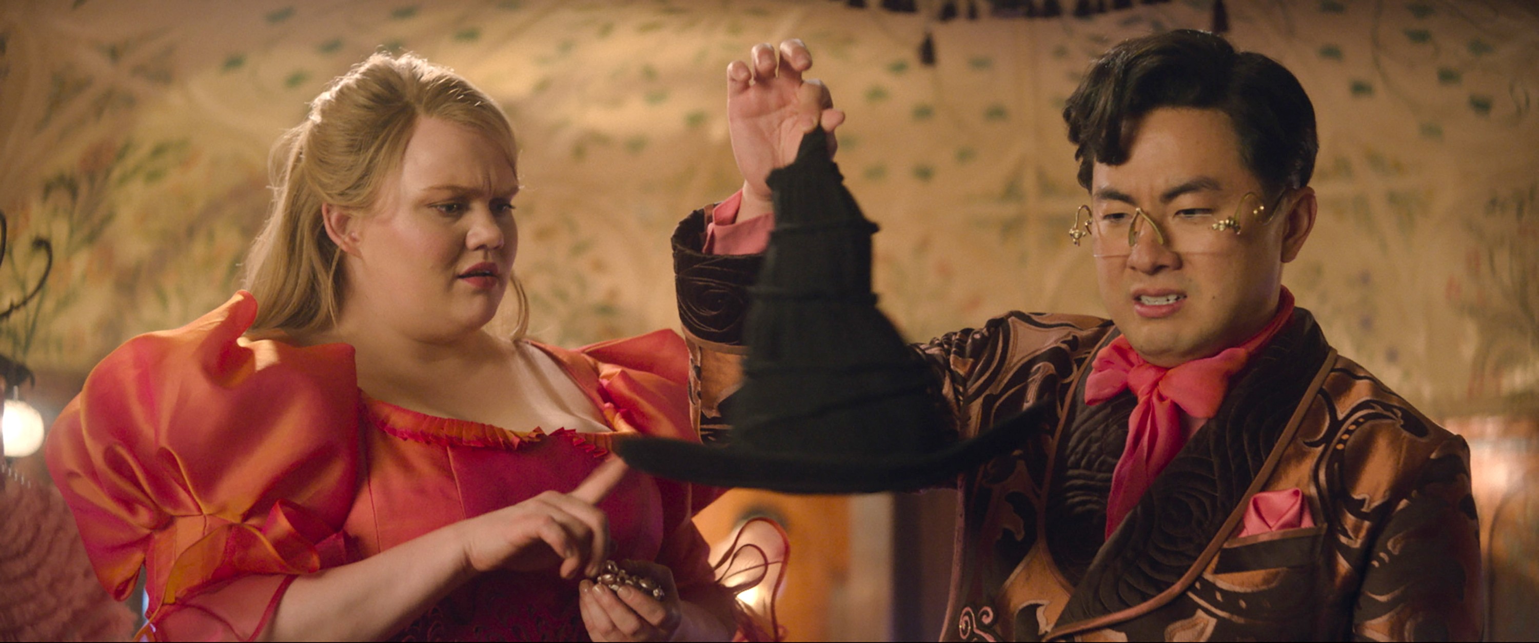 Two people in elaborate fantasy formalwear (Bronwyn James and Bowen Yang) hold up a pointed black witch’s hat and grimace at it in disgust in Wicked