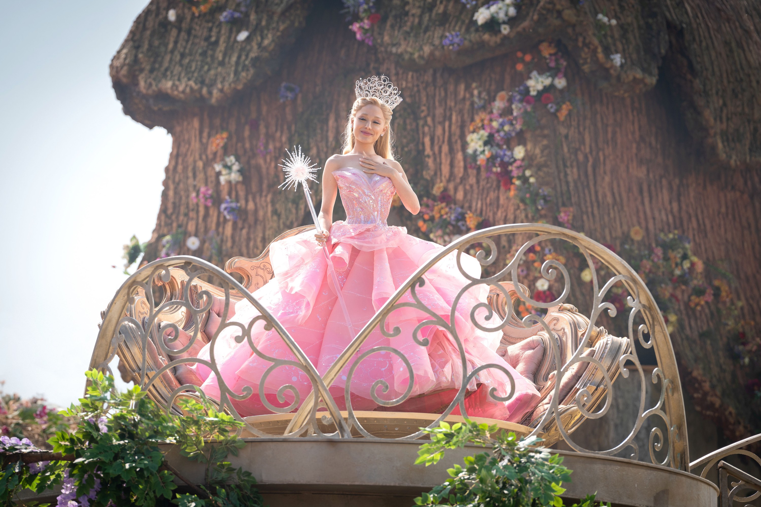 Glinda the Good (Ariana Grande, in a giant poofy pink ballgown, and carrying a wand with a sparkling silver-and-pink spangle on the tip) stands on a platform surrounded by metal lace balustrades and looks modest in Wicked