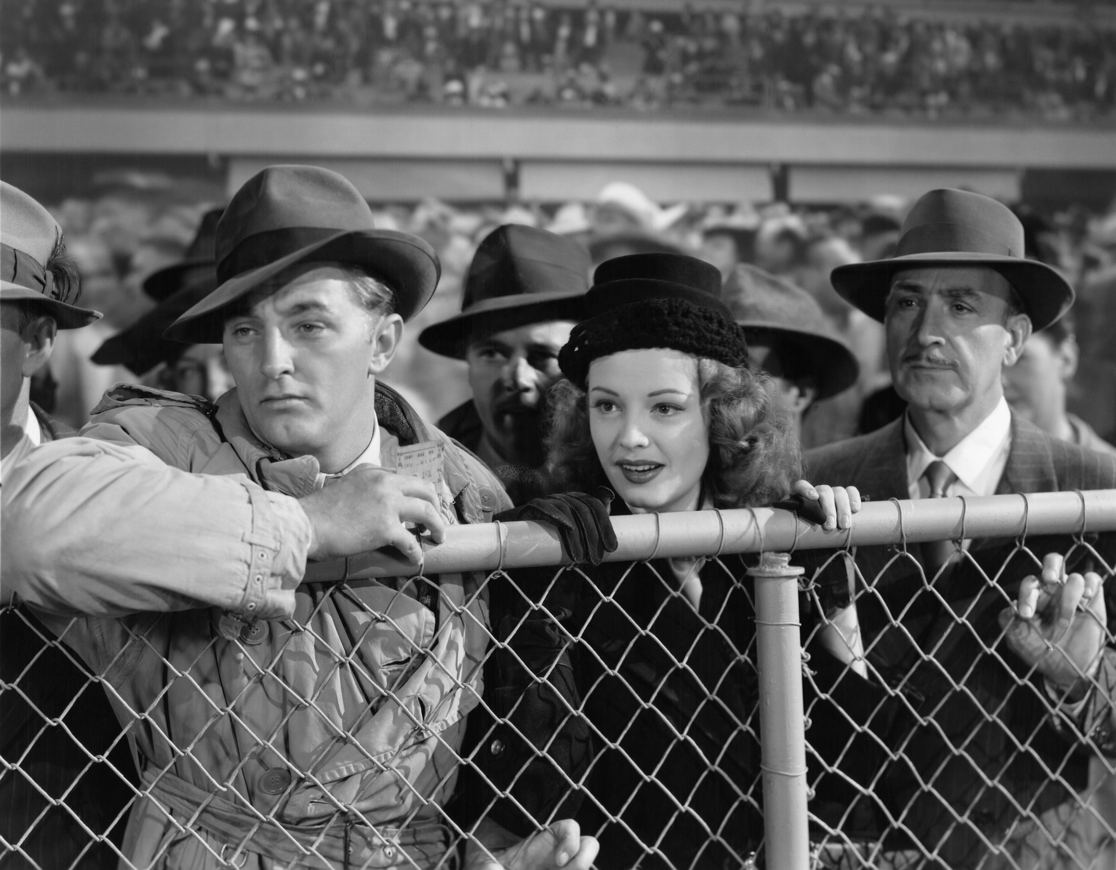 Robert Mitchum, Jane Greer, and a crowd look over a fence in Out of the Past