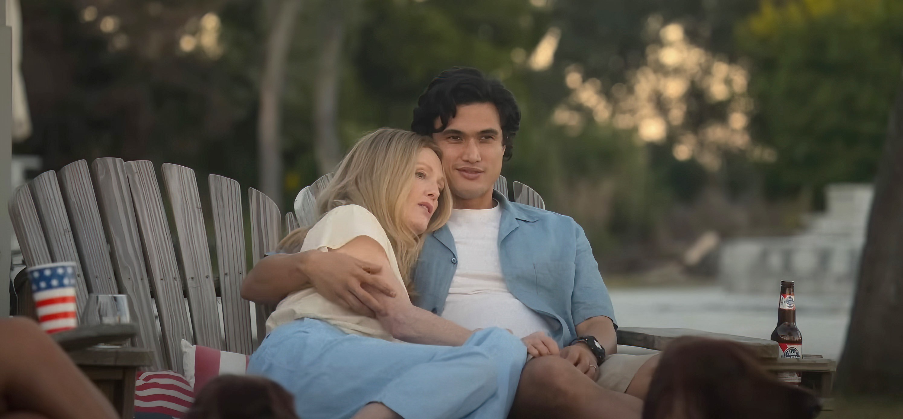 Joe (Charles Melton) and Gracie (Julianne Moore) together on a wooden outdoor bench on their lawn, her leaning against his shoulder, his arm around her, in May December