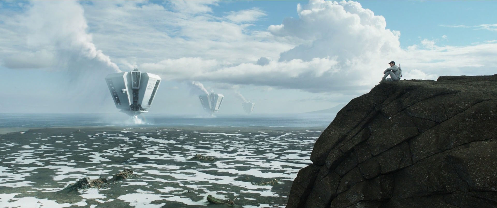 A man sits on the end of a cliff overlooking a vast landscape with gigantic floating machines sucking up water from the ocean in the distance.