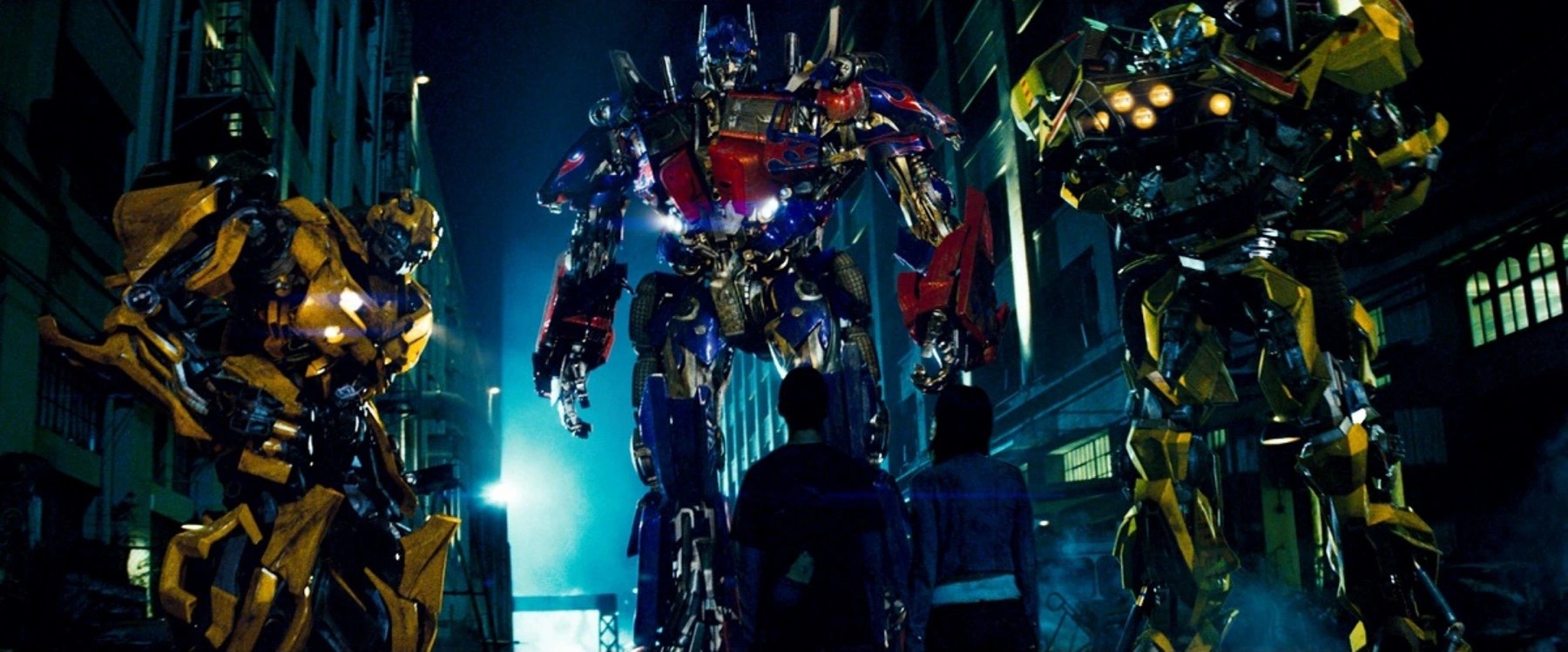 A trio of giant robots standing in front of a young man and woman in a darkly lit alley in Transformers