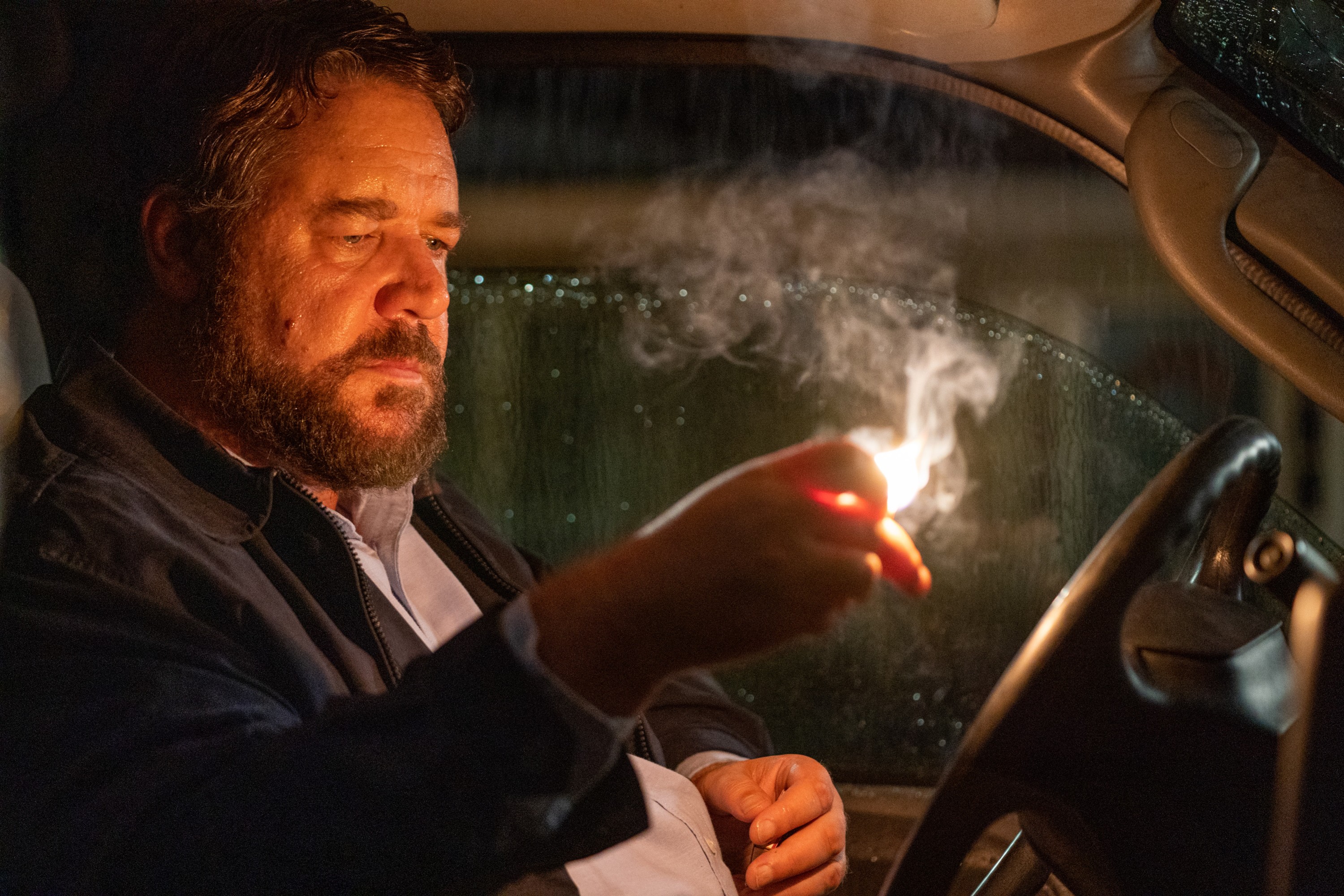 Russell Crowe sitting in the front of a pickup truck in Unhinged looking at a lit match in his hand