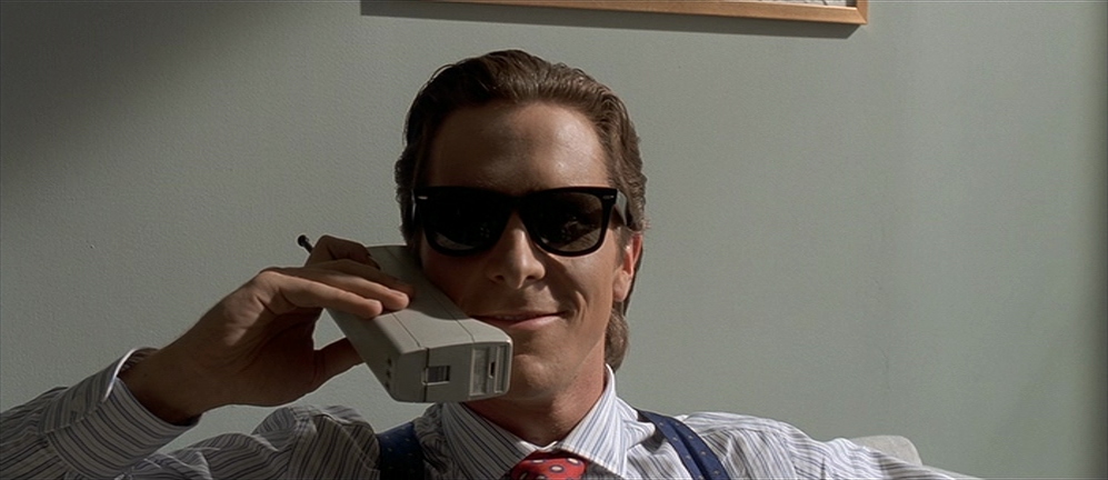 Christian Bale wearing sunglasses and talking into a large cellular phone in American Psycho.