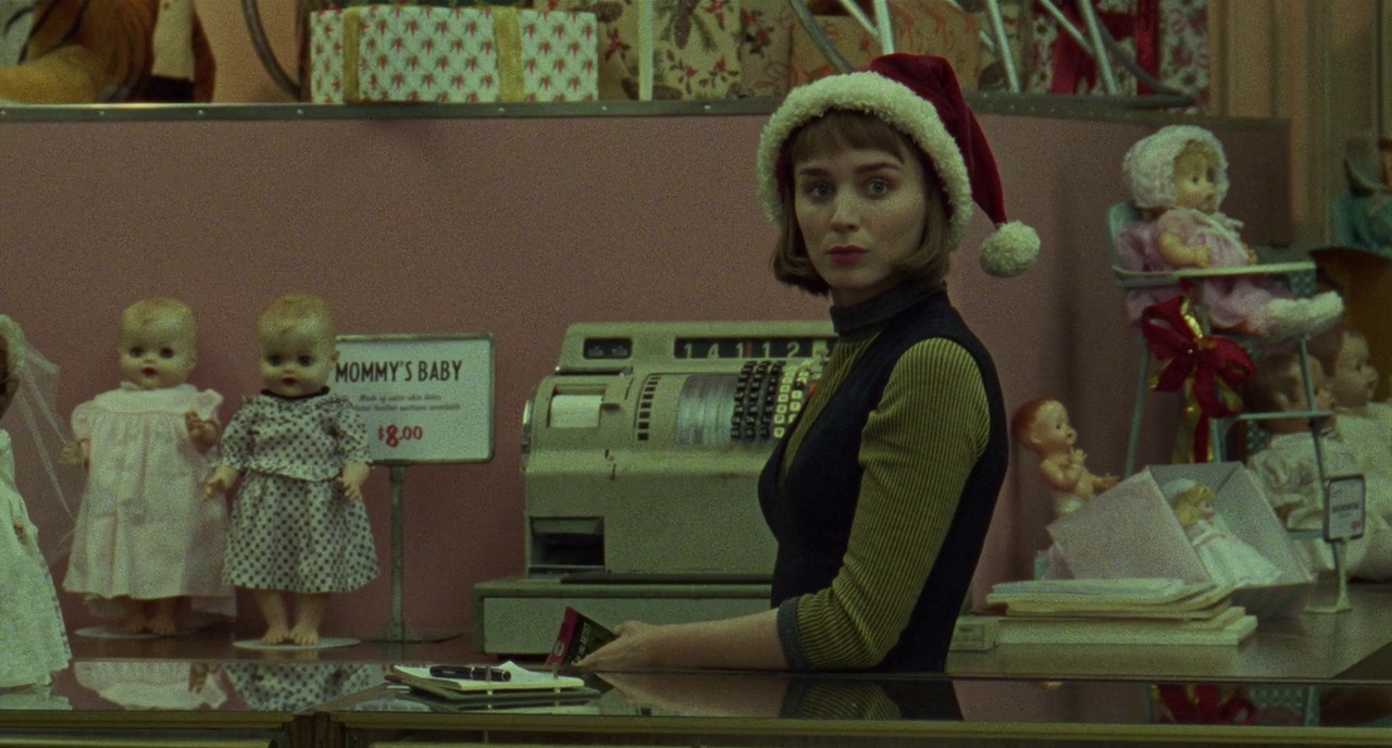 Rooney Mara wears a Santa hat behind a store counter. Baby dolls litter the background, and a sign reads “Mommy’s Baby.”