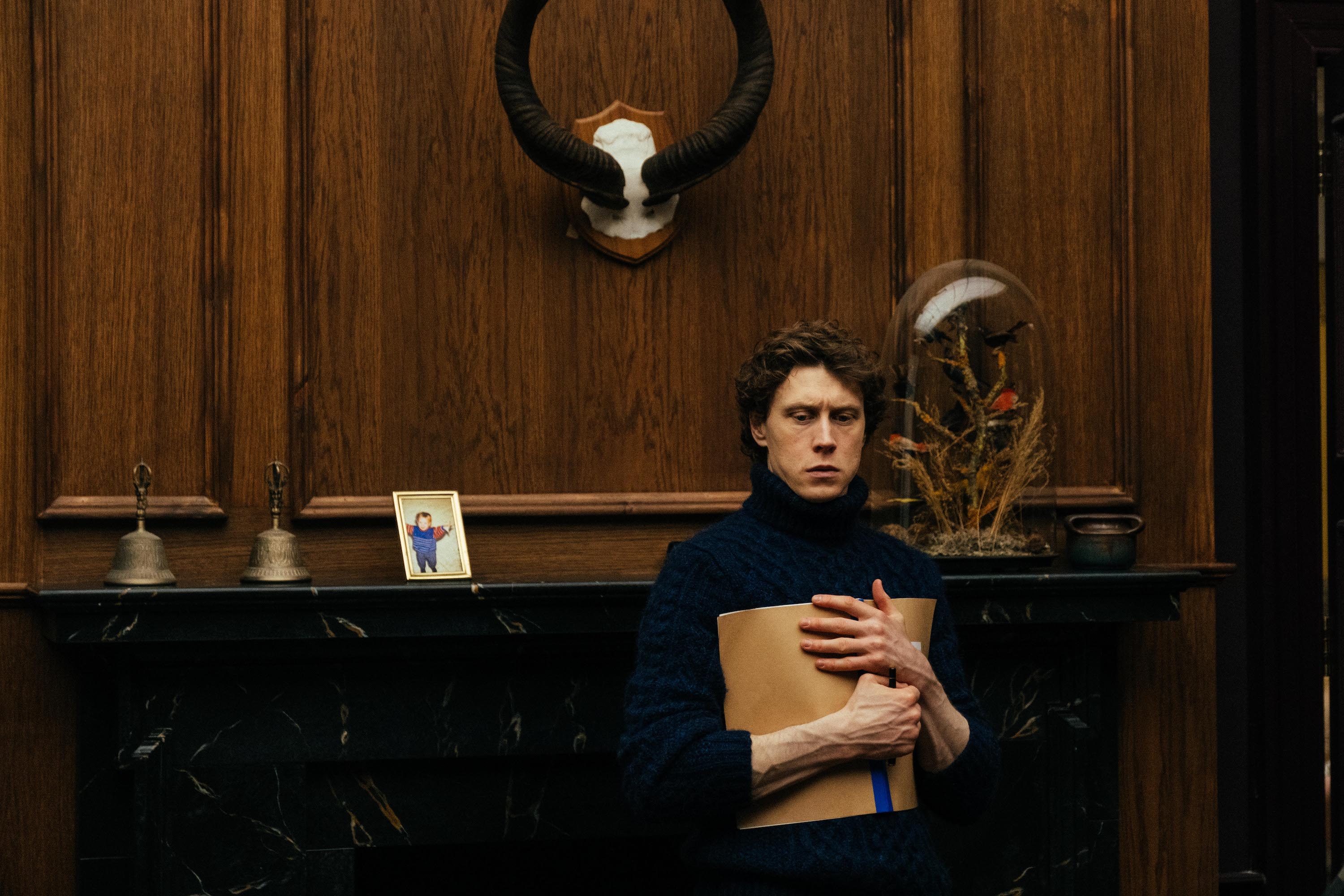 George MacKay, clutching a brown paper folder to his chest, stands in front of a wide fireplace surrounded by dark wood panelling in The End